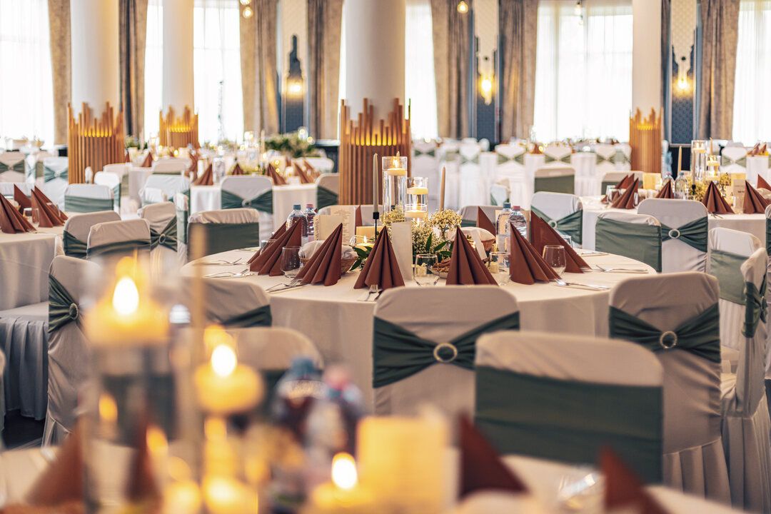 Candlelit banquet hall with round tables, white chair covers tied with green sashes and folded rust napkins