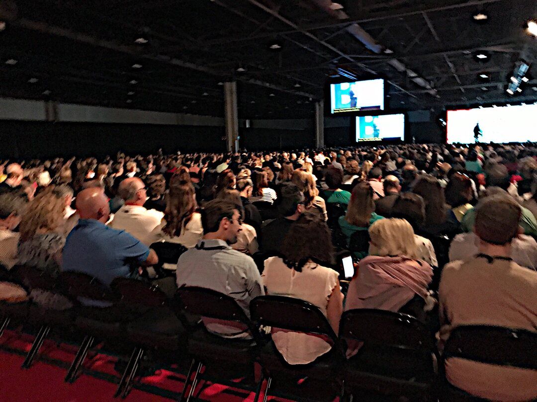 Seated conference audience facing a lit stage and large presentation screens in a dark hall