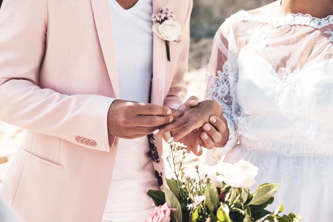 Groom in a pink suit sliding a ring onto the bride’s finger above a bouquet of white roses