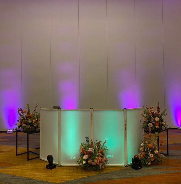 White DJ booth lit purple and teal, flanked by floral arrangements against a draped wall
