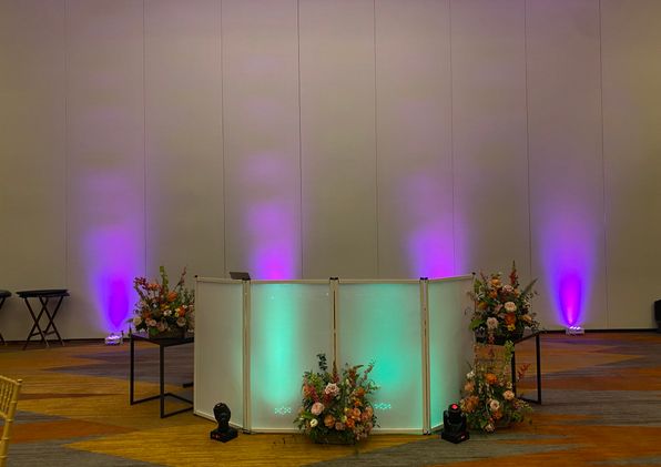 Purple uplights on a ballroom wall behind a glowing white DJ booth framed by flower arrangements