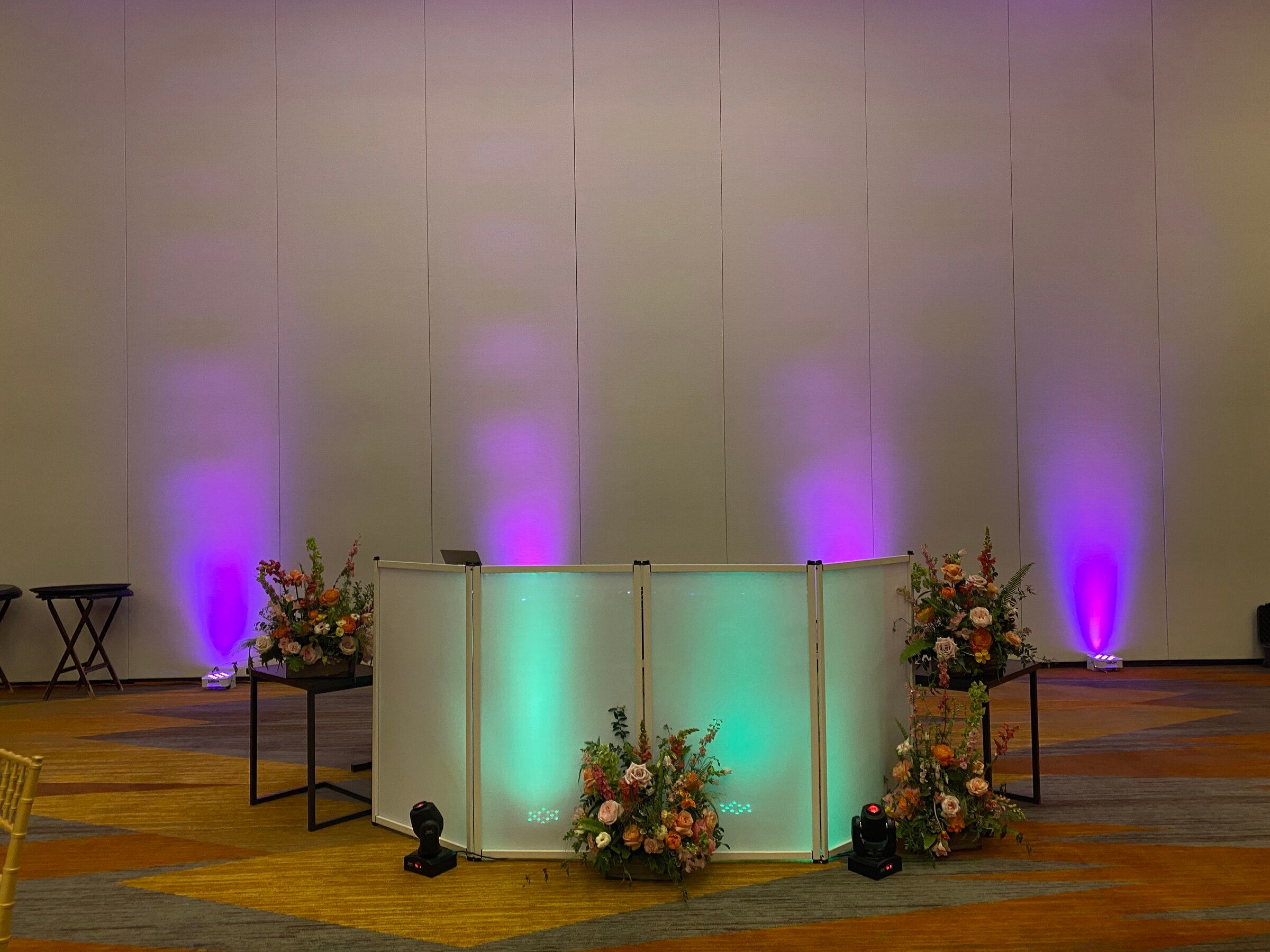 White DJ booth lit purple and teal, flanked by floral arrangements against a draped wall