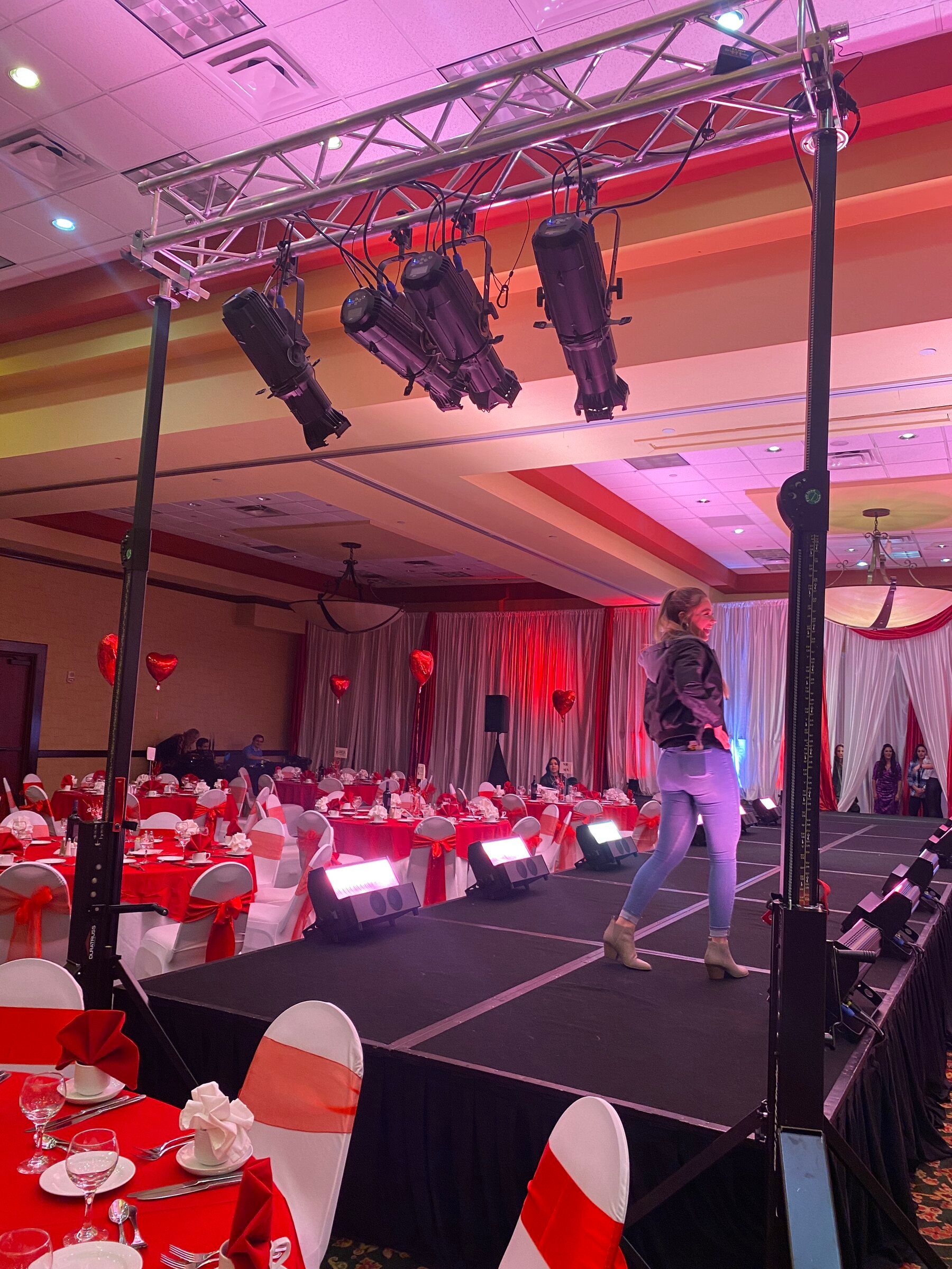 A model walking a banquet hall runway lit in pink and red by truss-mounted stage lights