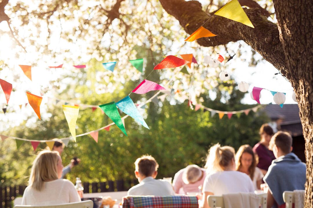 Guests seated at outdoor tables under a tree hung with colorful bunting and paper lanterns