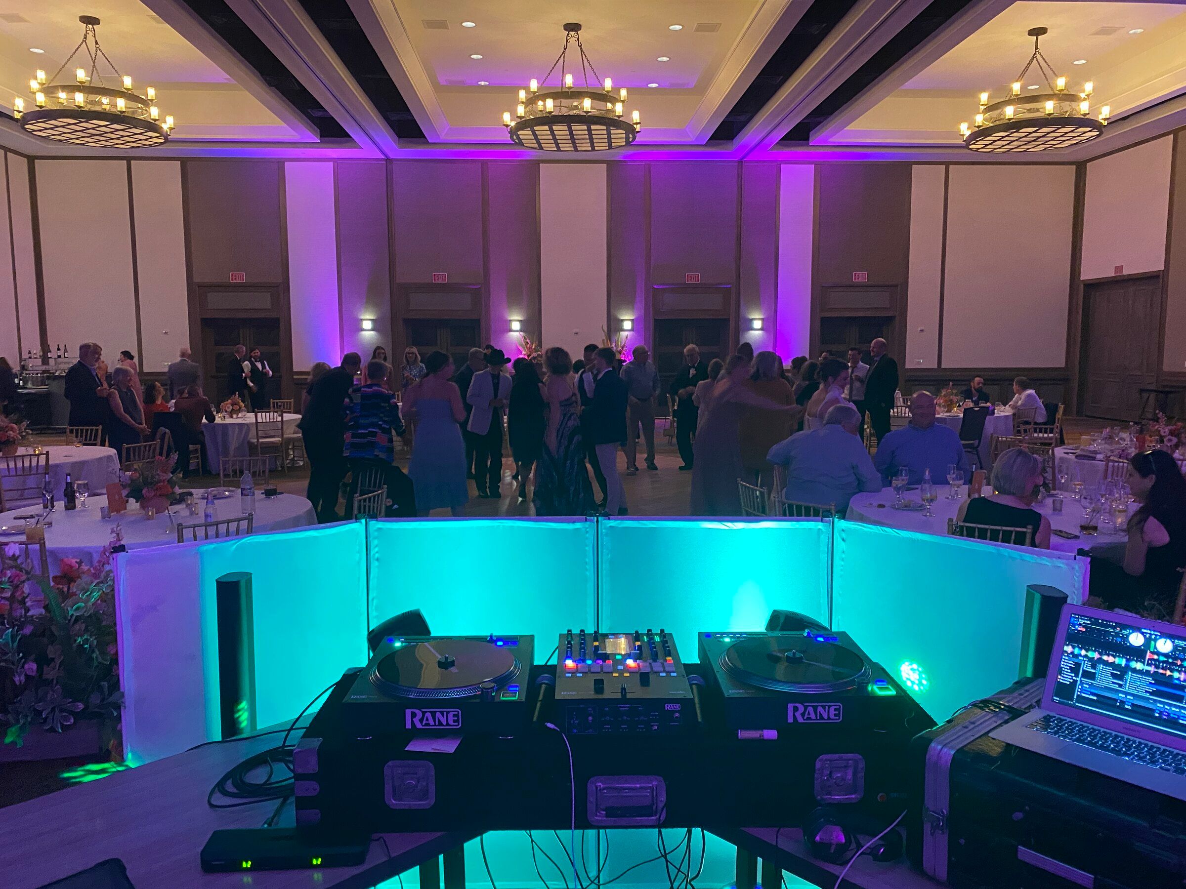 View from behind the turntables of guests dancing in a ballroom washed in purple uplighting