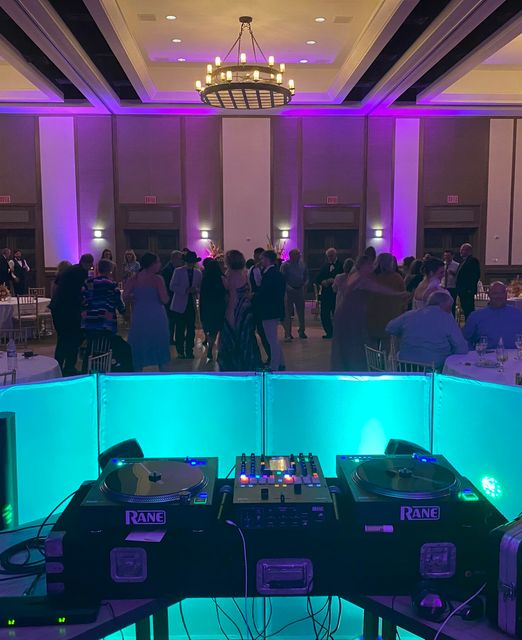 View from behind the turntables of guests dancing in a ballroom washed in purple uplighting