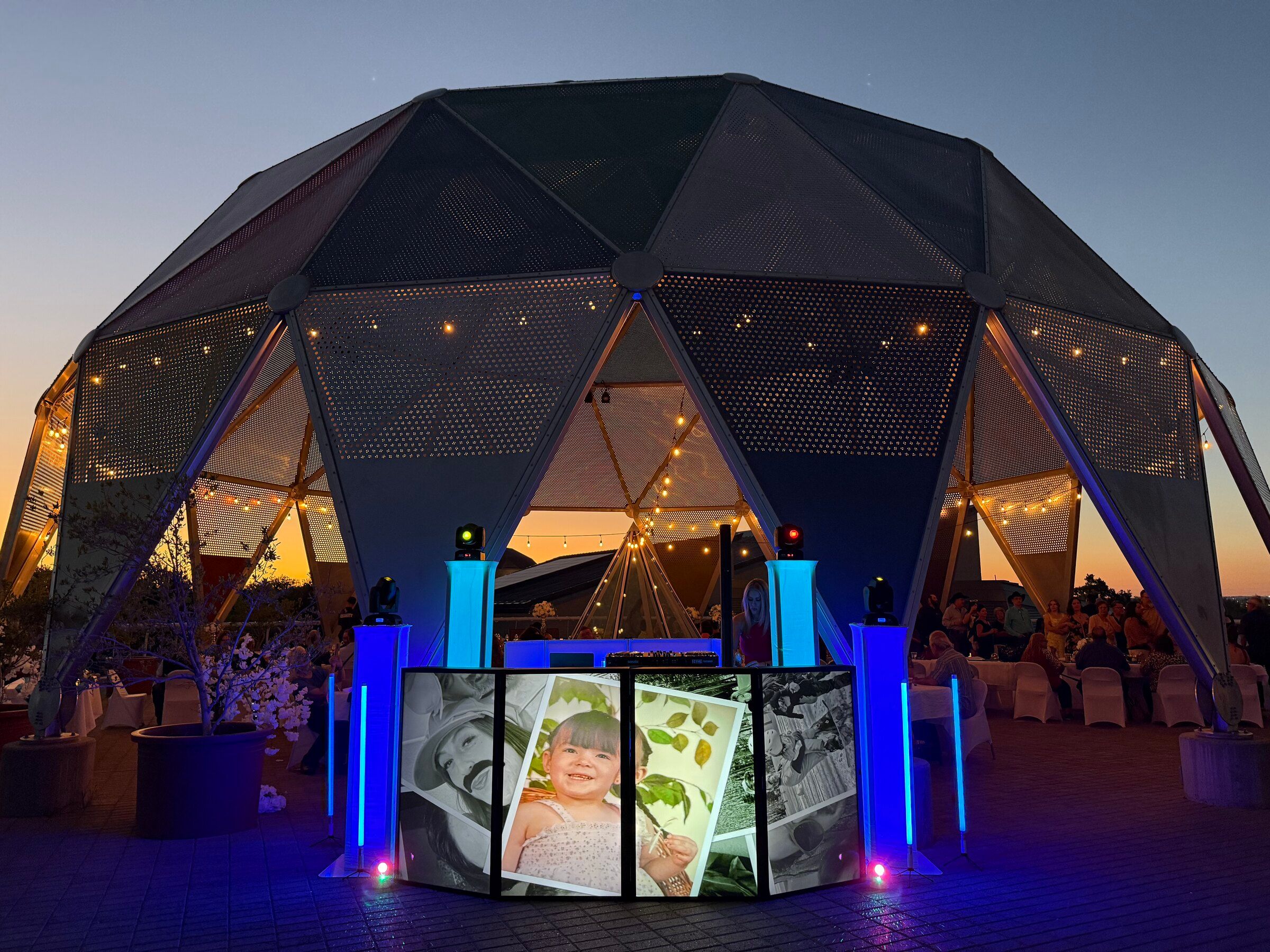 DJ booth with a photo display front and blue light towers outside a string-lit geodesic dome at sunset in Albuquerque