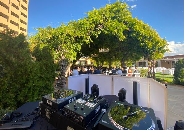 Turntables and mixer behind a white DJ booth facing a wedding ceremony under a vine-covered arbor