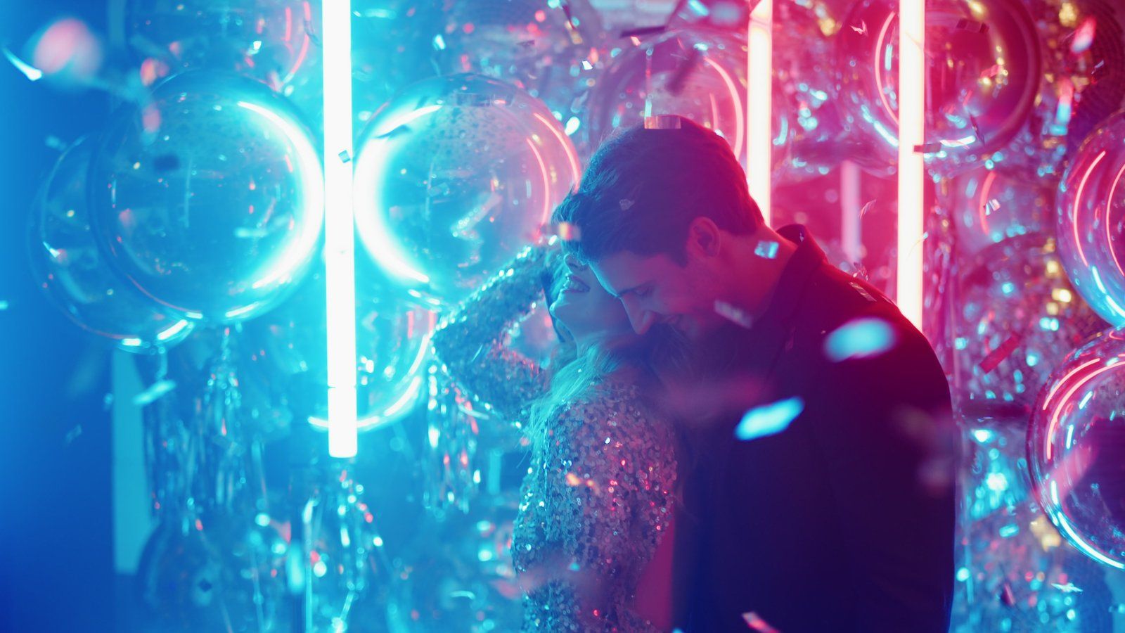 Couple embracing under falling confetti in a room lit with pink and blue neon and clear balloons