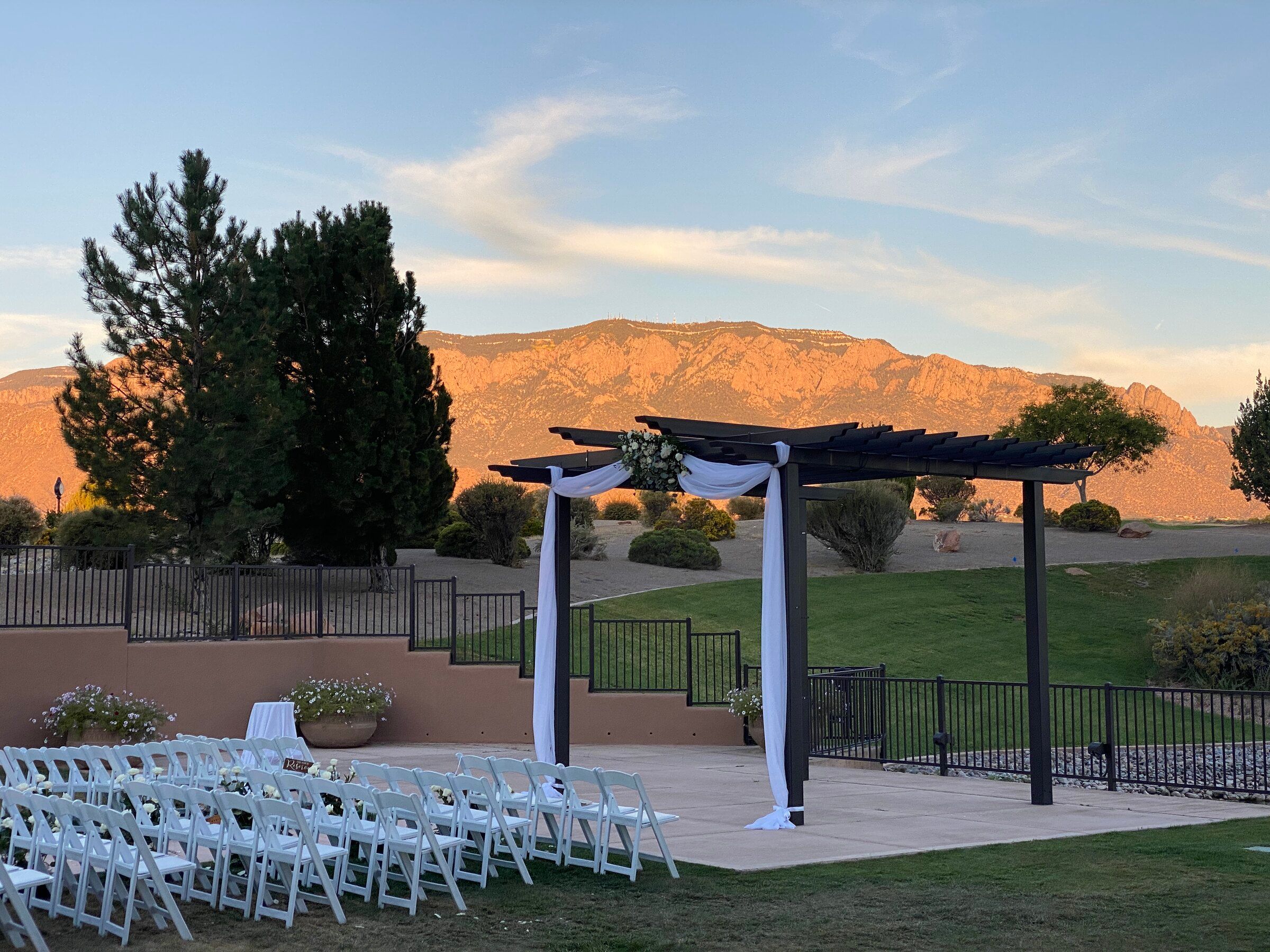 White chairs facing a draped pergola at an outdoor Albuquerque wedding ceremony with the Sandia Mountains glowing at sunset